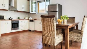 Eine Küche mit weißen Schränken, einem Edelstahlbackofen, einem Kühlschrank und Korbstühlen um einen Holztisch mit einer Obstschale.