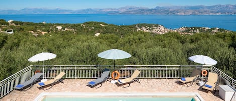 Ein Poolbereich mit Liegestühlen und Sonnenschirmen, mit freiem Blick auf das Meer und die dahinterliegenden Berge.