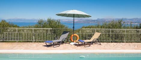Ein Poolbereich mit zwei Liegestühlen und einem Sonnenschirm, umgeben von einem Metallzaun und Blick auf ferne Berge.