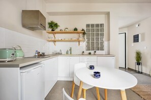Una cuina compacta amb armaris blancs, forns integrats i zona de dinar amb taula i cadires blanques.