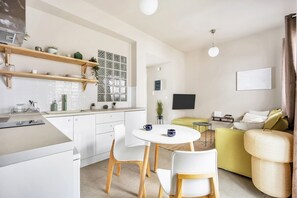 Una cuina moderna amb armaris blancs, prestatges de fusta i zona de menjador amb cadires blanques i una taula rodona. Hi ha un sofà i una televisió a la zona de sala d'estar.