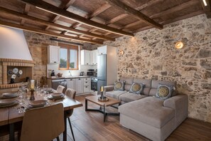 A rustic interior with a stone wall, wooden ceiling beams, a dining area with a table set for four, and a sofa with patterned cushions. - Stella's Traditional House Near the City (Vamvakopoulo)