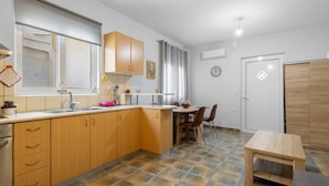 A modern kitchen with wooden cabinets, a dining area with a table and chairs, a small wooden stool, and a tiled floor. - Litsa’s Maisonette Near Sea - City Center (Chania)