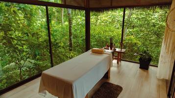 A room with a bed, a small table with a plant, and a view of dense greenery through large windows.