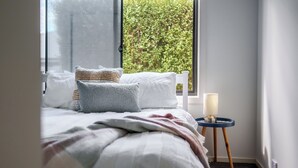 Et soveværelse med en seng, puder, et natbord med lampe og et vindue med udsigt til grønne omgivelser.