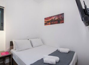 Et hotelværelse med en seng, hvide puder, et gråt sengetæppe og et billede af en by.