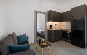 Een knus leefruimte met kitchenette, een bank met blauwgroene kussens, een klein tafeltje met croissants en een kopje, en een bed zichtbaar door een geopende deur.