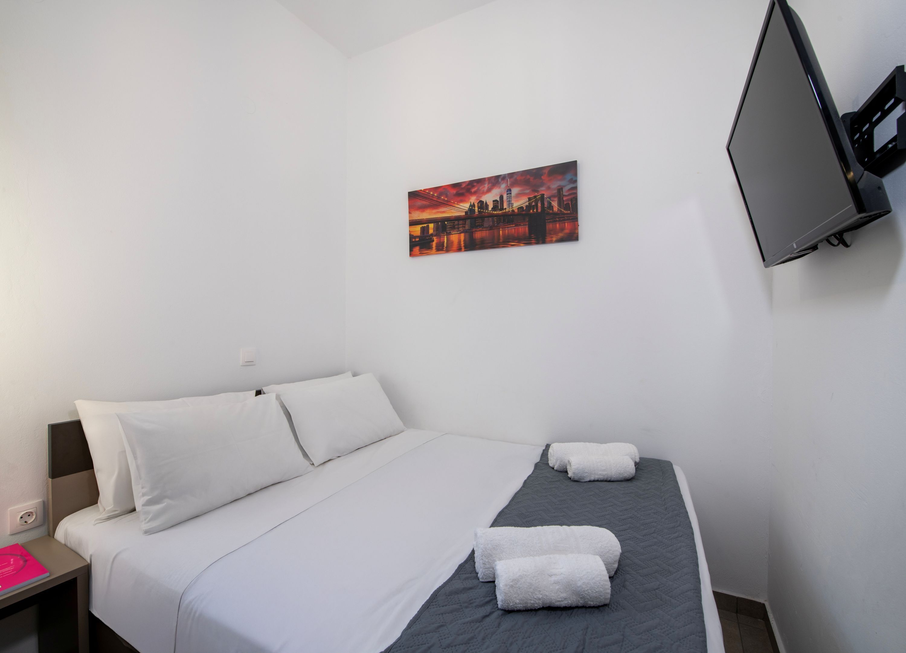 Une chambre d’hôtel avec un lit, un tableau représentant un paysage urbain, un téléviseur à écran plat et une table de chevet sur laquelle se trouve un objet rose.