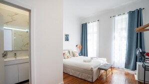 A modern bedroom with a bed, bedside table, and a small stool. There is a window with curtains, a mirror, and a wall-mounted fan. - Zarco Suites by Sé Funchal (Funchal)