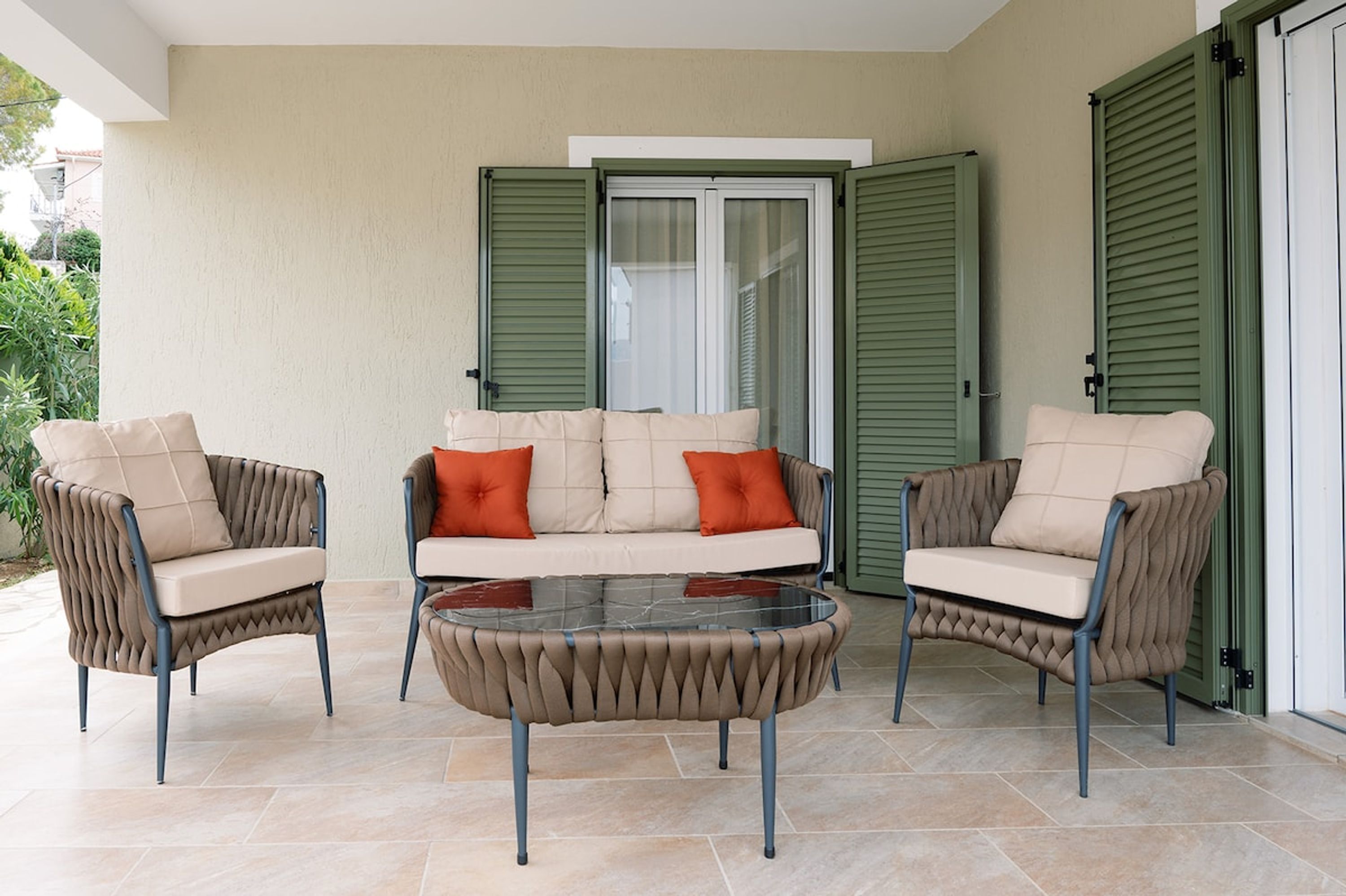 Eine Terrassenfläche mit einer Couch und zwei Sesseln, einem Glastisch und einer Schiebetür mit grünen Fensterläden.