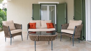 Een terras met een bank en twee fauteuils, een glazen salontafel, en een schuifdeur met groene luiken.