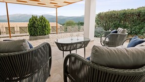 Un espace extérieur aménagé, abrité et doté de mobilier en osier et d’une table en verre, offrant une vue sur un paysage montagneux.