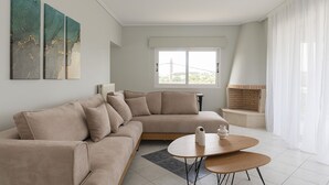 Moderni olohuone, jossa on beige värinen kulmasohva, pieni pyöreä pöytä ja ikkuna, jossa on läpikuultavat verhot.