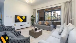Un salon moderne avec un canapé gris, un canapé blanc et une table basse. Un téléviseur à écran plat est fixé au mur, et une grande fenêtre est agrémentée de rideaux.
