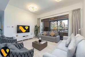 Ein modernes Wohnzimmer mit grauem Sofa, weißem Sofa und Couchtisch. An der Wand ist ein Flachbildfernseher befestigt und ein großes Fenster mit Vorhängen vorhanden.
