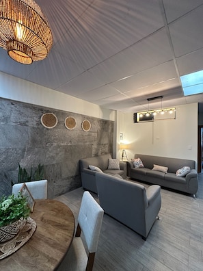 Ein modernes Wohnzimmer mit einer grauen Sofagarnitur, einem Holzespeisetisch mit Stühlen und einer großen, gewebten Leuchte.