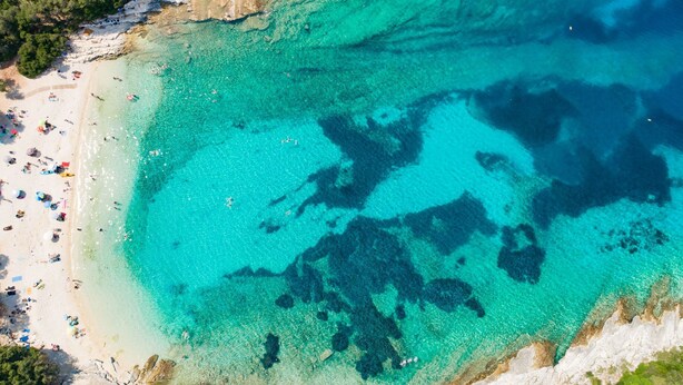 Ett flygbildsbild av en strand med kristallklart turkost vatten, en klippig kustlinje och en sandstrand med parasoller och människor.