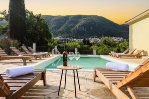 Zona de piscina con sillas de descanso, una mesa con una botella y vasos, y vista a un pueblo y montañas al fondo.