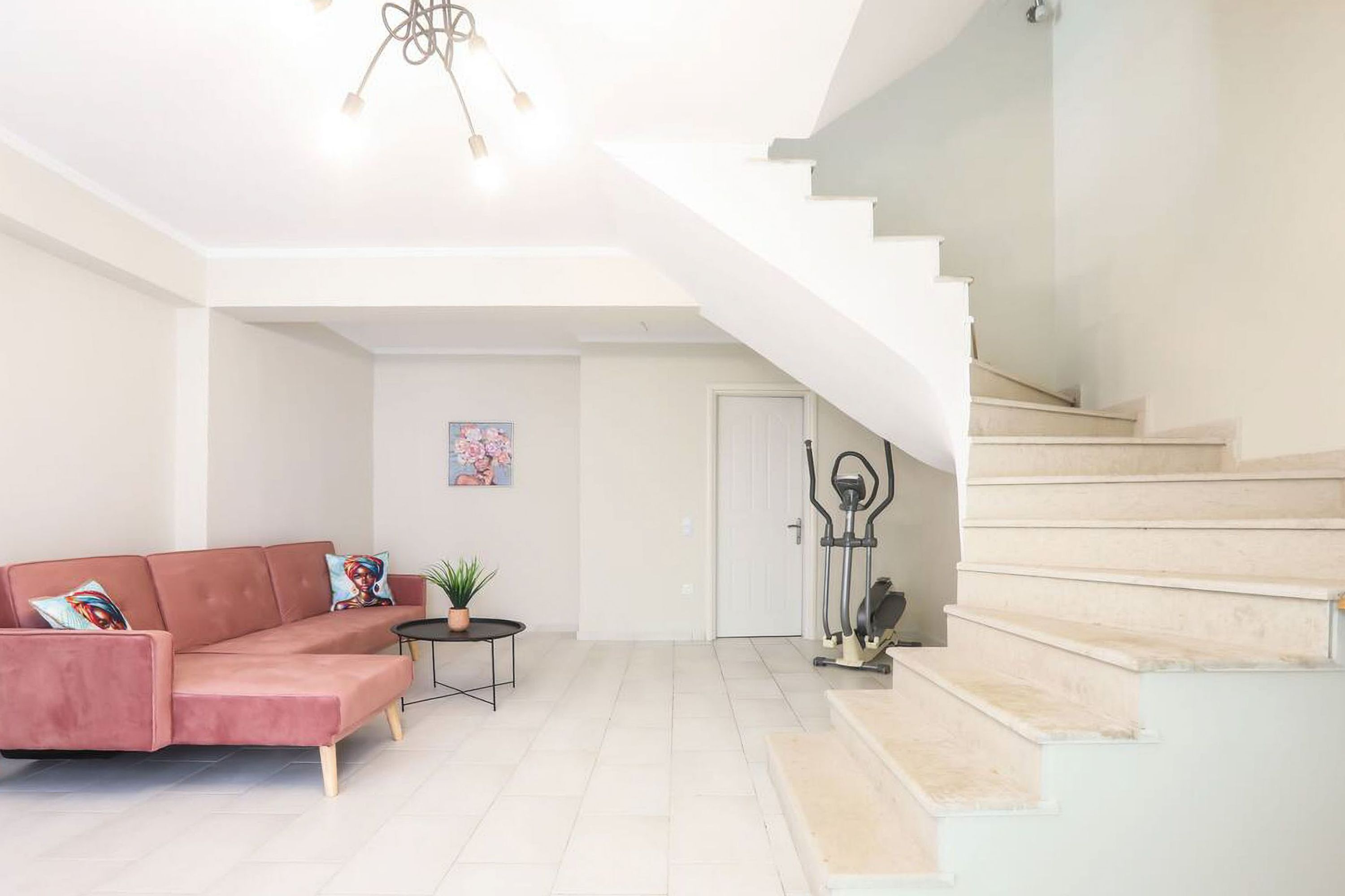Un salon spacieux avec un canapé rose, une petite table ronde et une plante en pot. Un escalier mène à l’étage supérieur.