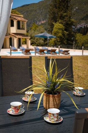 A table with floral cups and saucers, a potted plant, and a view of a pool and lounge chairs. - Elite Villa Dio in Ano Korakiana (Ano Korakiana)