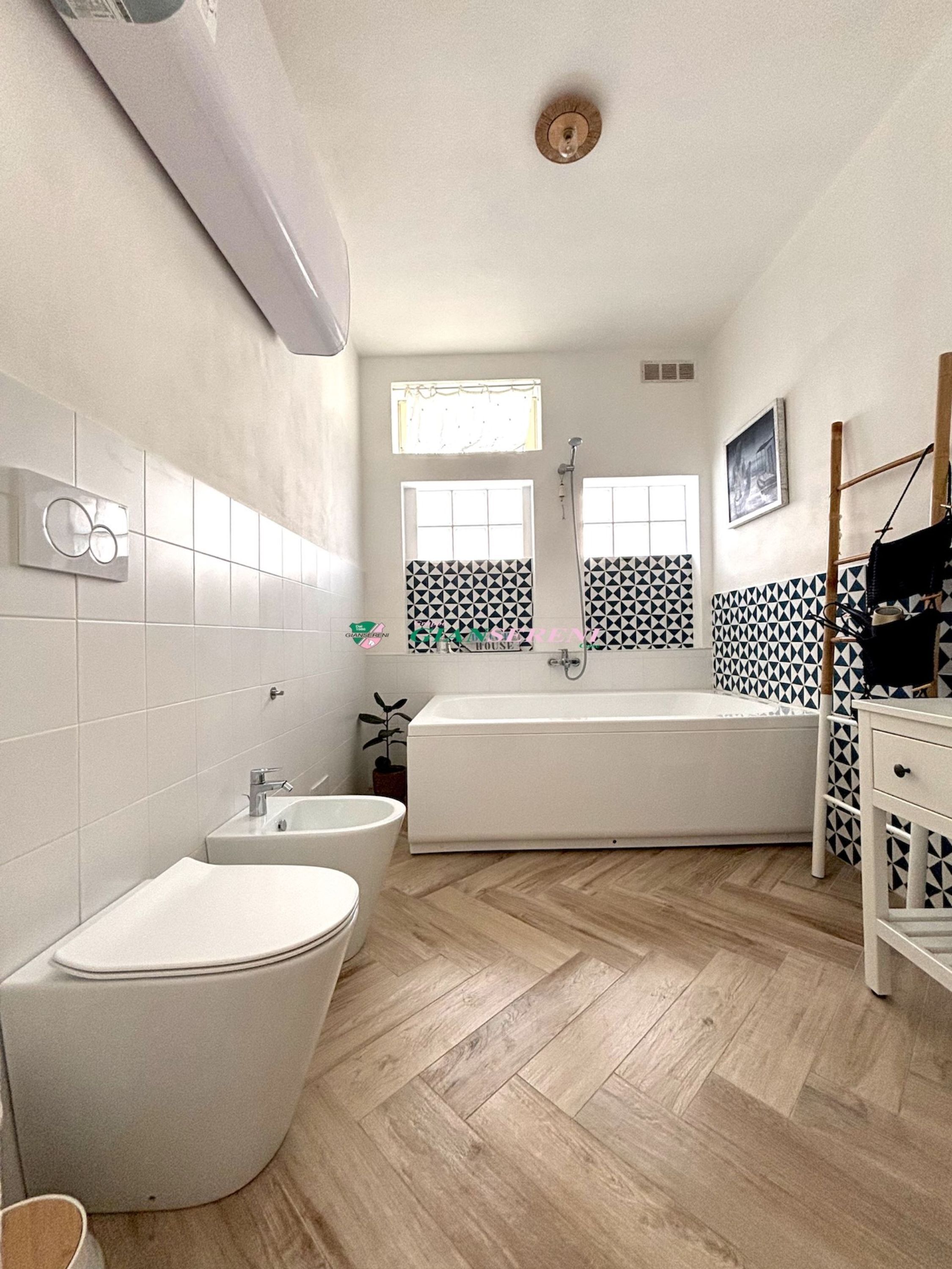 Une salle de bain moderne avec une baignoire sur pieds, des toilettes et un lavabo. Deux fenêtres laissent entrer la lumière naturelle, et le sol est recouvert de parquet disposé en motif chevrons.