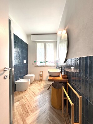 Une salle de bain moderne avec un parquet en bois disposé en chevrons, deux bidets blancs, un grand miroir et un tabouret en bois.