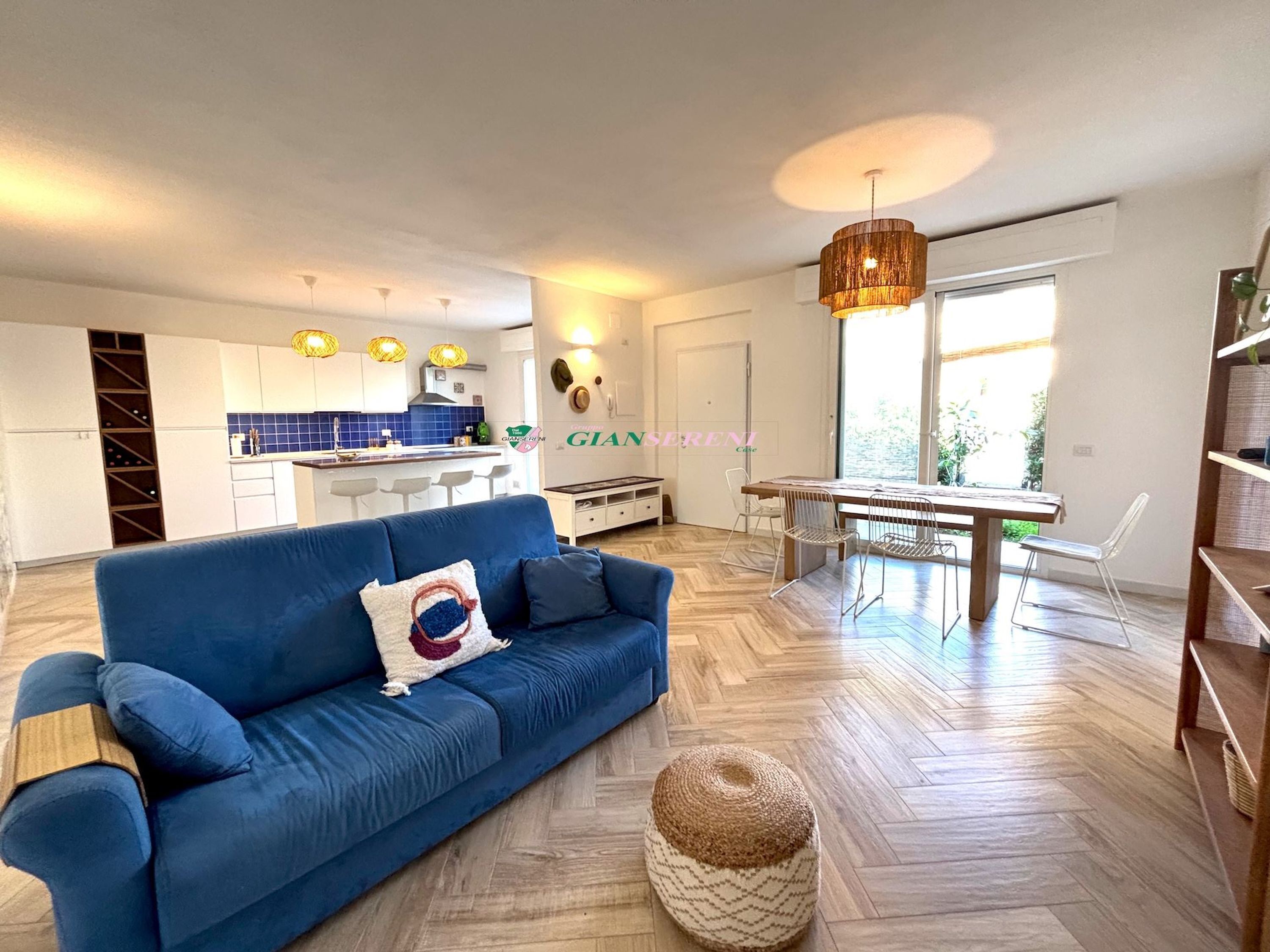 En moderne stue med en blå sofa, et spisebord med stole, et køkken i baggrunden og et stort vindue.