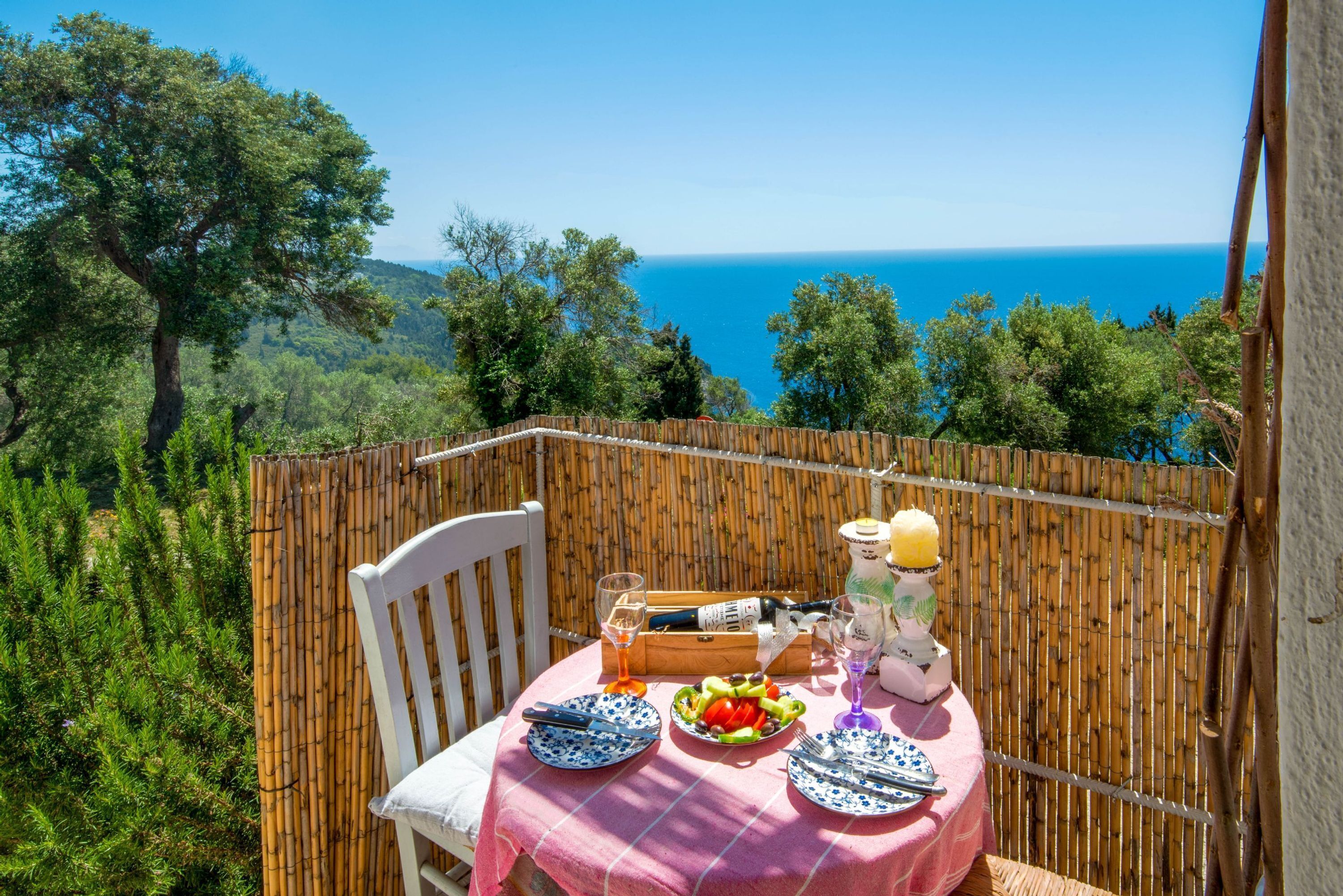 Ein Tisch ist mit Tellern, Besteck, einer Flasche und Gläsern gedeckt und bietet einen schönen Blick auf das Meer und Bäume.