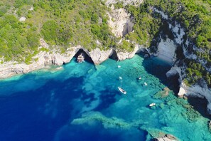 An aerial view of a coastal area with clear turquoise waters, rocky cliffs, and a boat anchored near a cave entrance. - Thodoroulis in Paxi with 1 bedrooms and 1 bathrooms (Paxi)