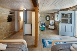 Ein gemütliches Zimmer mit Steinwand, Holzboden, einem Bett mit blau-weiß gemustertem Teppich, einem kleinen Tisch mit Stuhl und einer Tür mit Metallgitter.