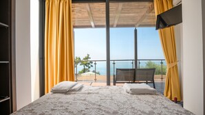 Ein Schlafzimmer mit Bett, zwei Handtüchern, einem Sessel und Meerblick durch große Fenster.