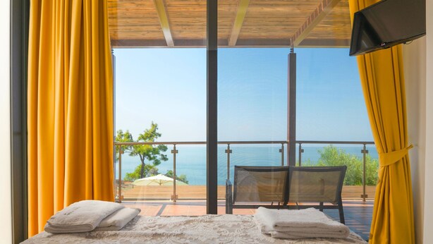 Ein Zimmer mit einem Bett, einem Sessel und einem Fernseher. Es bietet Meerblick und einen Balkon mit gelben Vorhängen.