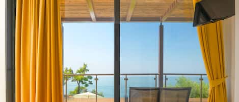 A room with a bed, a chair, and a TV. It has a view of the sea and a balcony with yellow curtains.