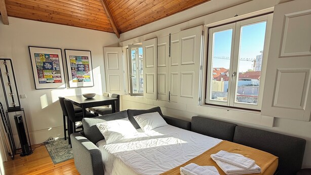 A bedroom with a wooden ceiling, a bed with a white and brown bedspread, a gray sofa, a desk with a chair, and a window with a view of buildings. - Charming apartment in the center of Porto! Lots of sunshine and a garage available. (Porto)