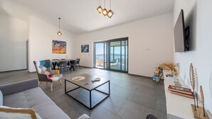 Ein geräumiges Wohnzimmer mit grauem Sofa, Couchtisch und Essbereich mit Tisch und Stühlen. Ein großes Fenster bietet einen Blick ins Freie.
