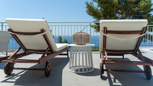 Dos sillones de exterior con ruedas en un balcón, una mesita redonda con una cesta de mimbre, y vistas al mar y a los árboles.