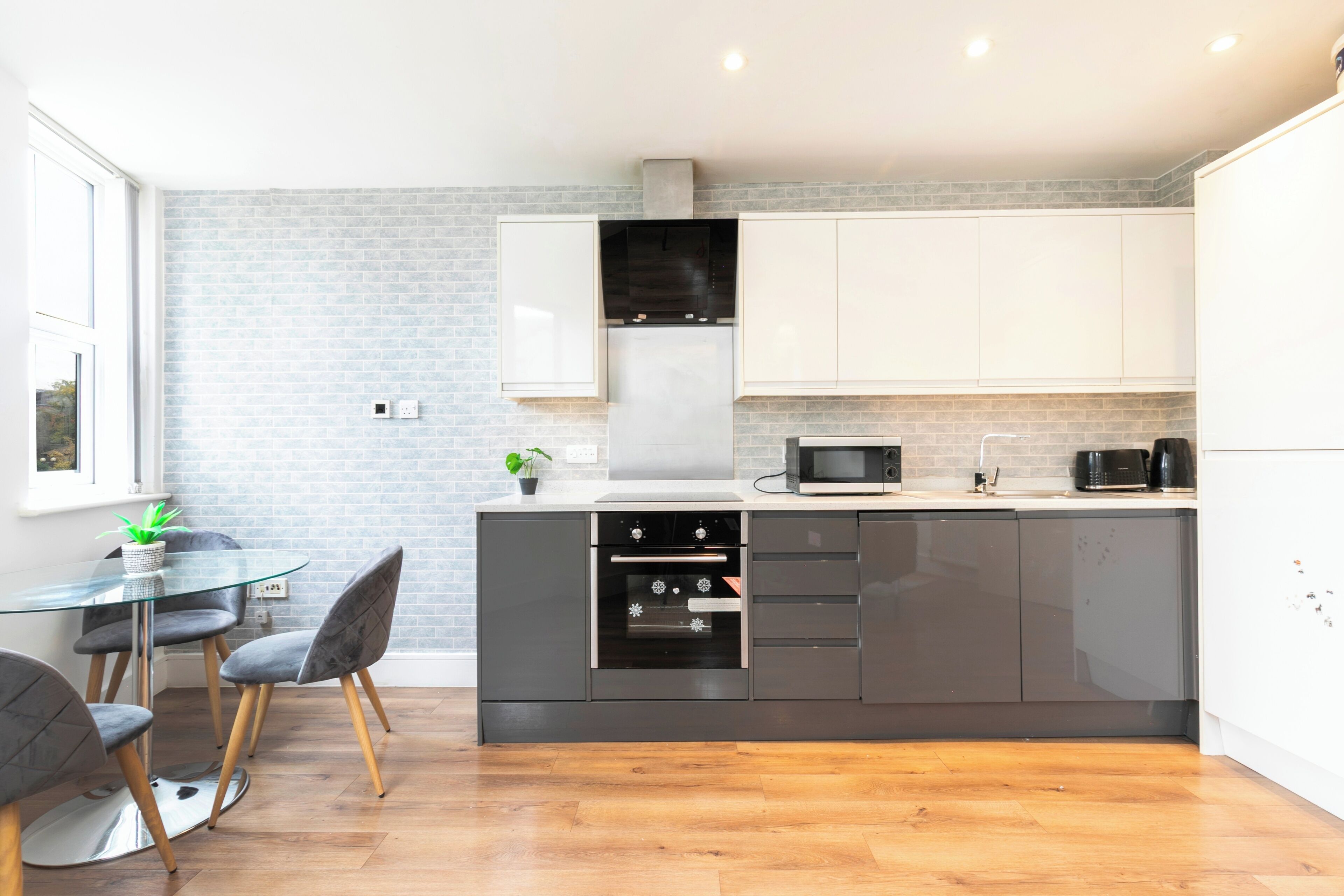 A modern kitchen with a glass dining table, grey chairs, and a brick wall.