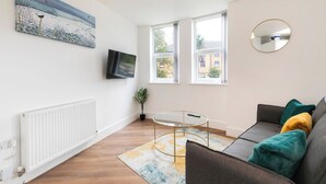 Ett modernt vardagsrum med en soffa, ett runt glasbord, en tavla, en tv och stora fönster.
