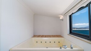 Ein modernes Badezimmer mit einer großen Badewanne, Holzboden, einem Fenster mit Meerblick und einem kleinen Ablagebrett mit einer Lampe.