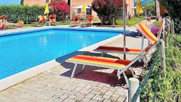 En pool med solstolar och parasoller, omgiven av en innergård med tegelbeläggning och grönska.