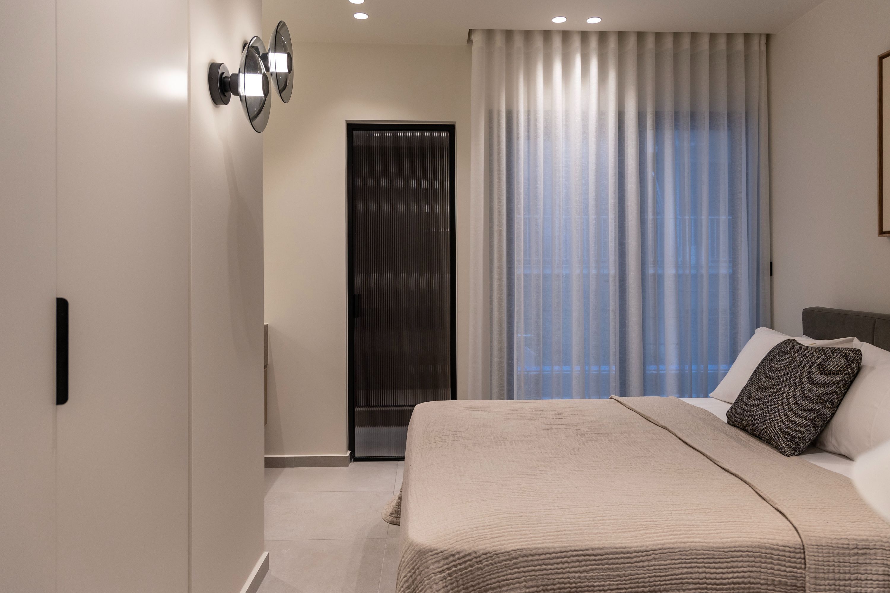 A modern bedroom with a large bed, a door with a vertical handle, and a window with sheer curtains.
