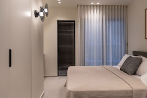 A modern hotel room with a large bed, a door with a black handle, and a window with sheer curtains.