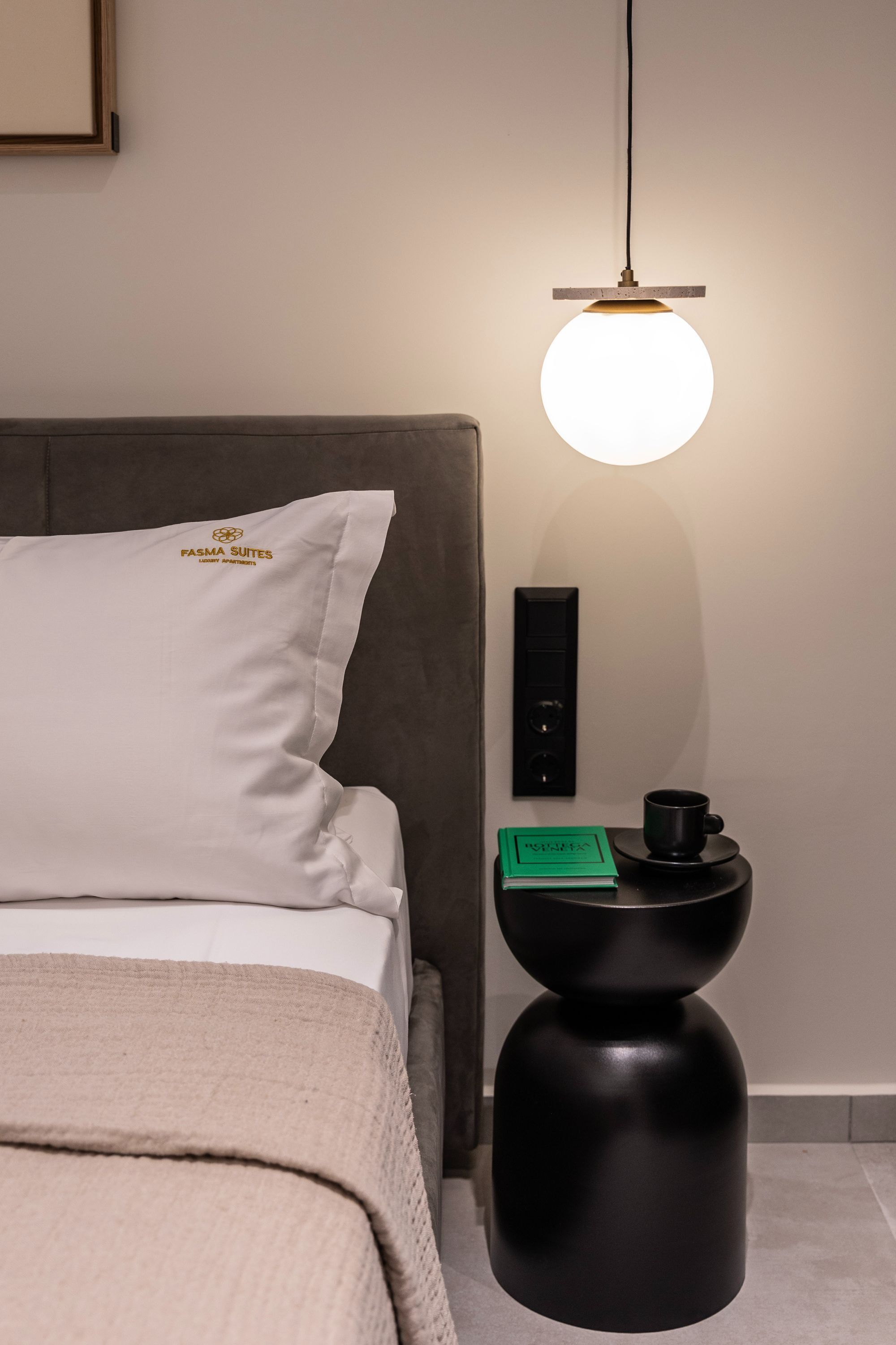 Un lit bien fait, avec un oreiller portant l’inscription « FASMA SUITES », et une table de chevet sur laquelle se trouvent une lampe noire, une boîte verte et une tasse noire.