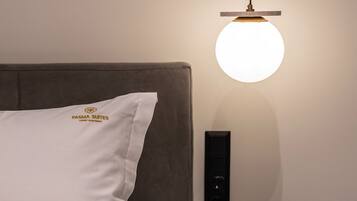 Un lit bien fait, avec un oreiller portant l’inscription « FASMA SUITES », et une table de chevet sur laquelle se trouvent une lampe noire, une boîte verte et une tasse noire.