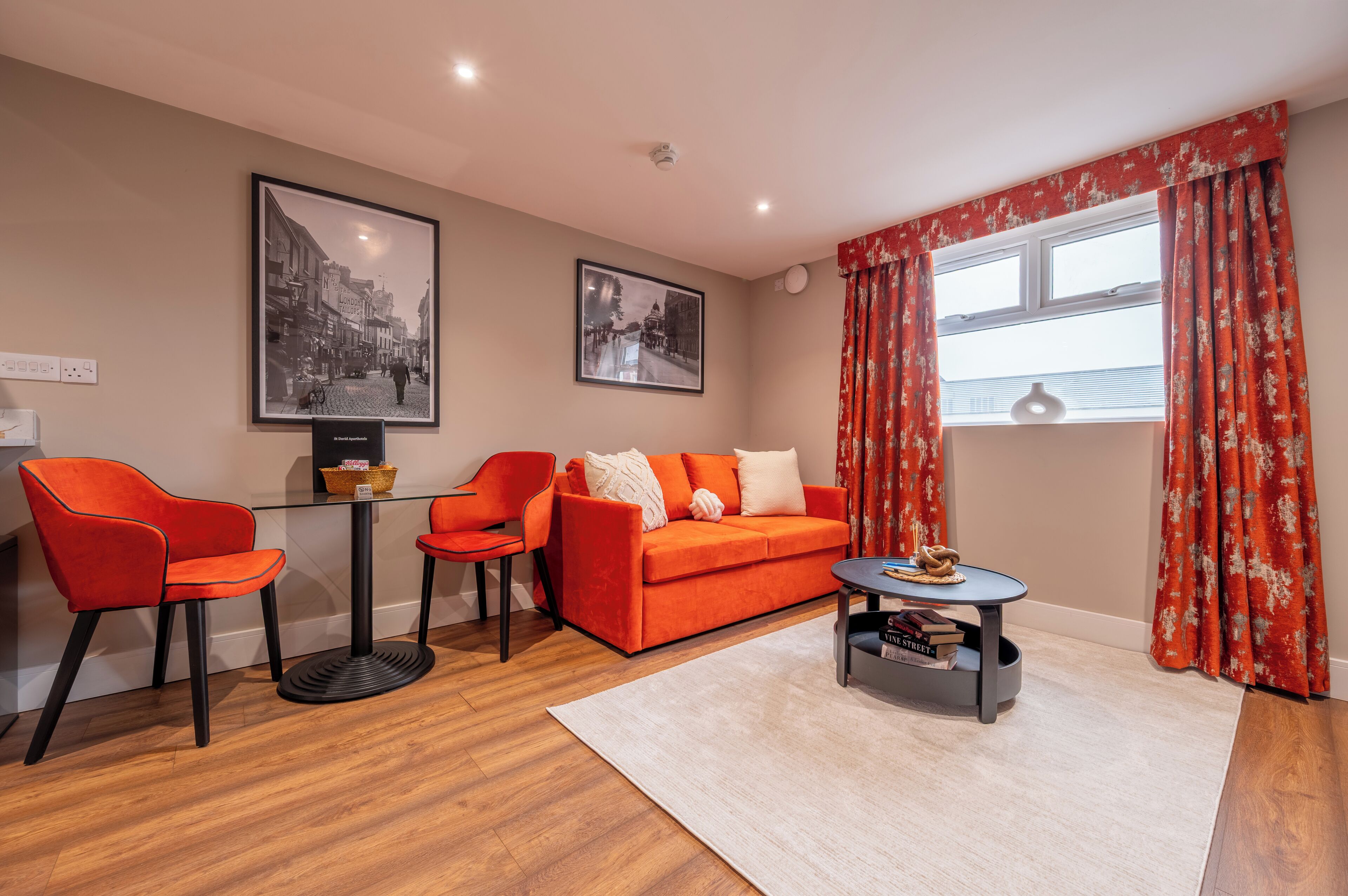 Un salon moderne avec un canapé, deux fauteuils, une table basse et une petite table d’appoint. Deux tableaux encadrés ornent le mur, ainsi qu’une fenêtre avec des rideaux rouges et un interrupteur.