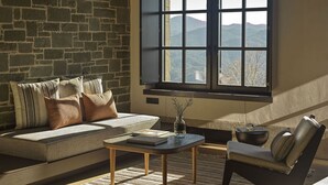 A cozy living room with a stone wall, a large window with a mountain view, a sofa, a chair, and a small table with books and a vase.