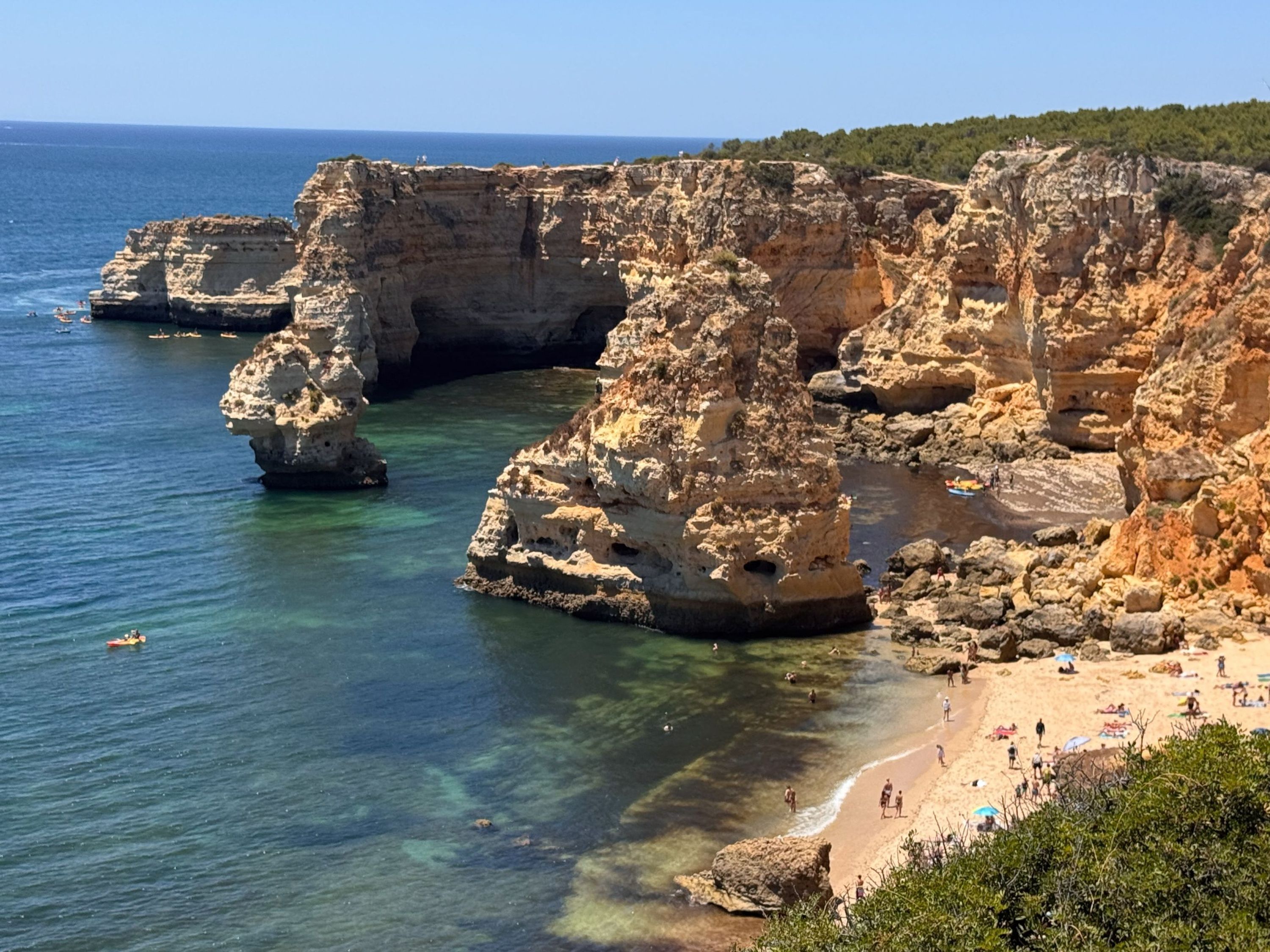 A coastal landscape with rocky cliffs, clear blue sea, and a sandy beach with people.