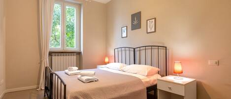 A bedroom with a bed, two bedside tables with lamps, a window with curtains, and framed pictures on the wall.