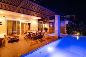 A modern outdoor pool area with a wooden deck, a dining table with chairs, and a glass-enclosed patio. - Sunny Exclusive Dio in Chaniotis (Chaniotis)