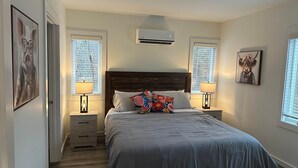 Un dormitorio con cama, dos mesitas de noche con lámparas, aire acondicionado de pared y dos ventanas con persianas.
